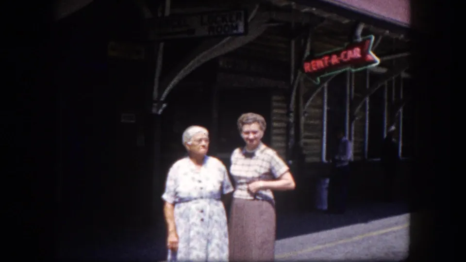 Two elderly women pose outside what appears to be Tampa's train depot, circa 1960; a neon RENT-A-CAR arrow sign and arched facade visible behind them.