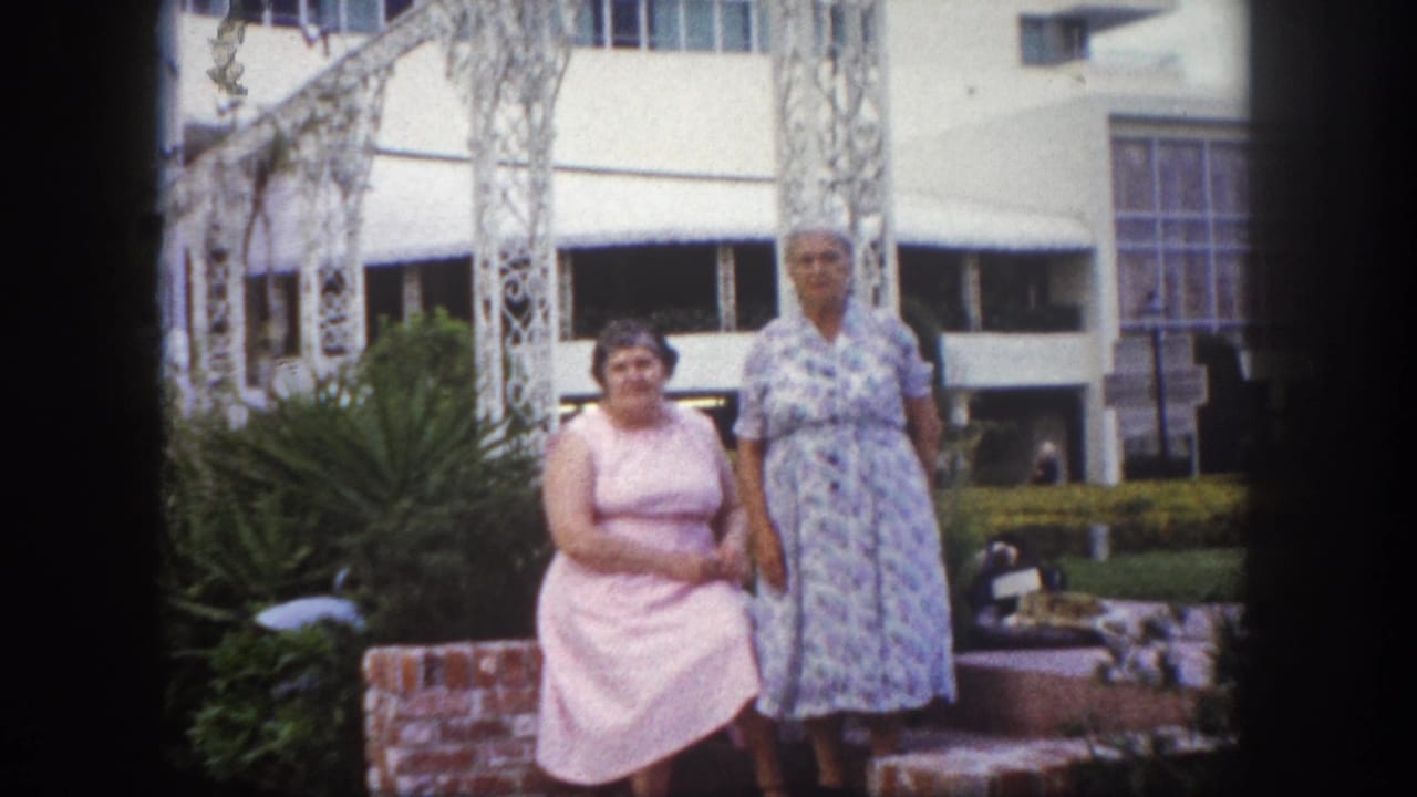 Two women in pink and blue floral dresses posing before ornate white metal arches and a mid-century modern building, Tampa, Florida, circa 1960, 8mm film.