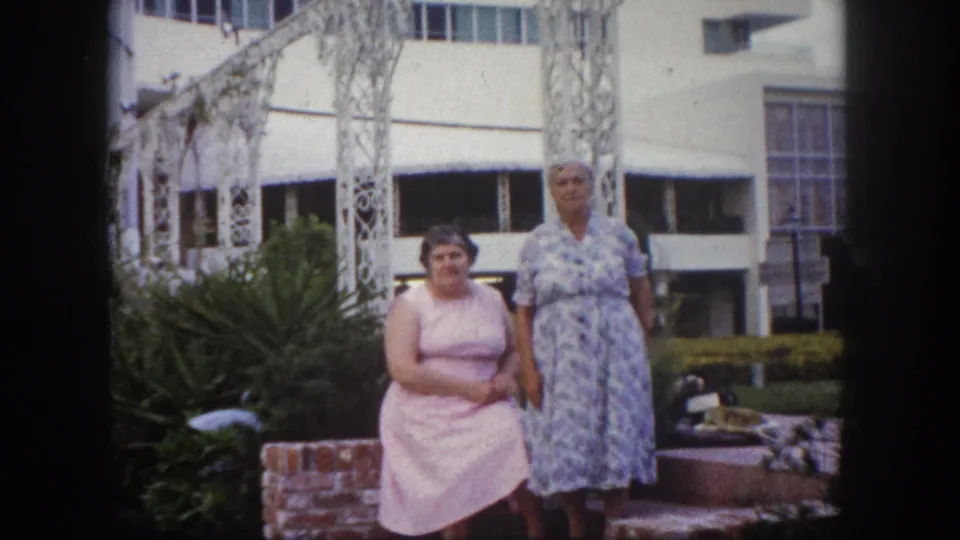 Two women standing together before white ornate metalwork and a mid-century modern building exterior, Tampa, Florida, circa 1960, 8mm film.