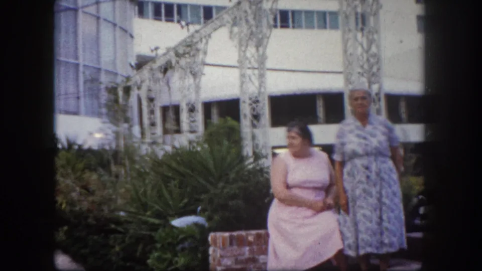 Two women posing before ornate white metal arches and a mid-century building exterior, Tampa, Florida, circa 1960, 8mm film.