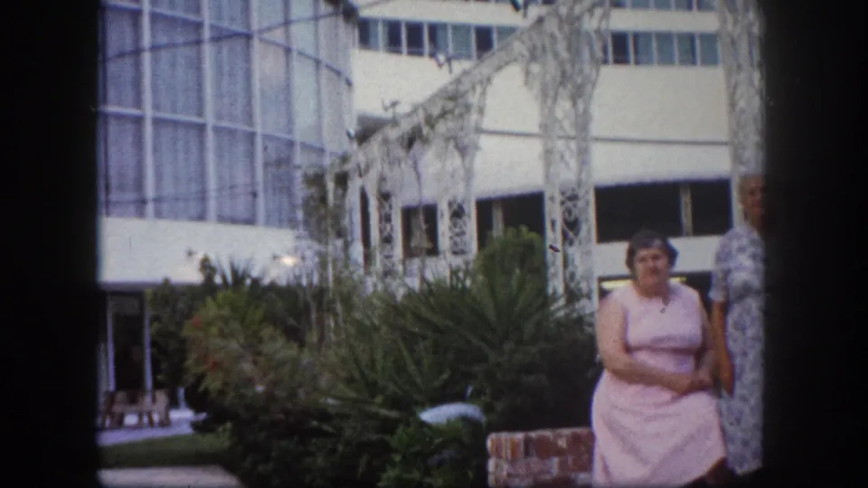 Woman in pink dress stands in foreground with ornate white metal pergola and mid-century building, Tampa, Florida, circa 1960, 8mm film.