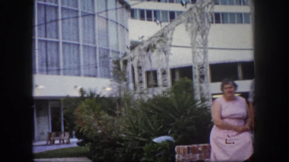 Woman in pink dress posing in a courtyard, likely Tampa Florida circa 1960, before a mid-century modern glass-facade building with white wrought-iron pergola and palm trees.