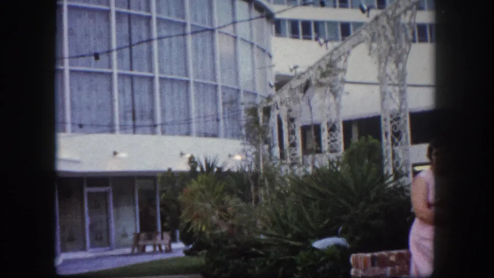 Mid-century modern glass curtain-wall building and white decorative pergola, likely Tampa Florida circa 1960; second woman in floral dress partially visible at frame right.