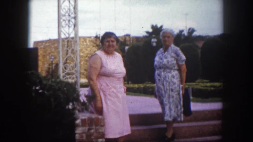 Two adult women posing together by a white ornamental metal trellis in an outdoor courtyard, likely Tampa Florida circa 1960, with palm trees and manicured hedges visible.