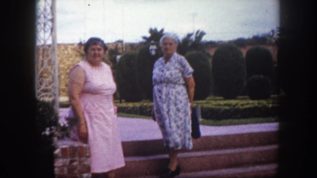 Two middle-aged women in 1960s-style dresses, one pink and one blue floral, stand near ornate metal garden structures and trimmed hedges, Tampa, Florida, circa 1960.