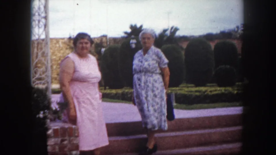 Two middle-aged women in 1960s-style dresses, one pink and one blue floral, stand near ornate metal garden structures and trimmed hedges, Tampa, Florida, circa 1960.