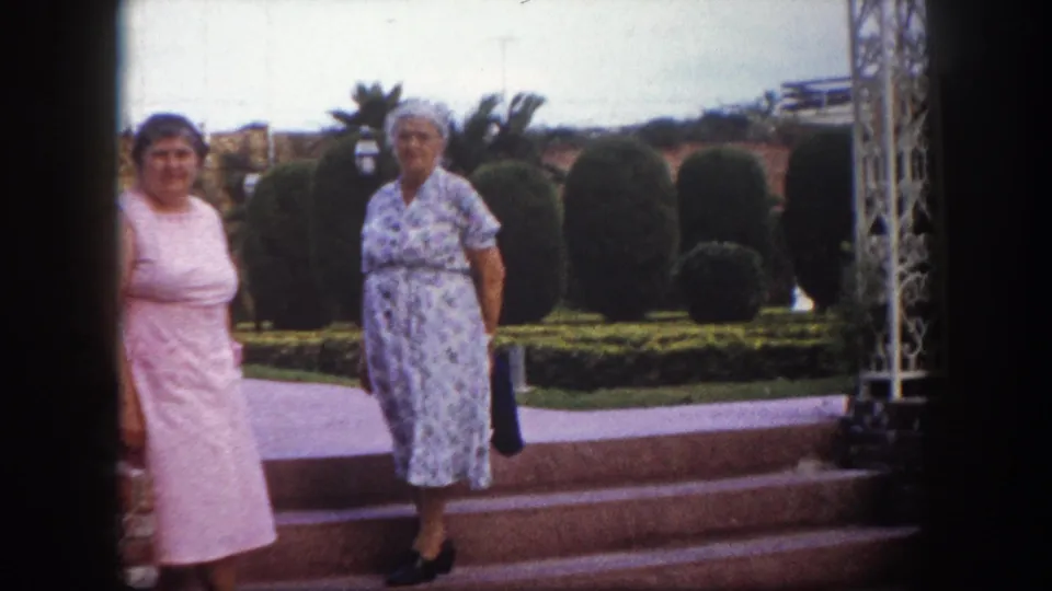 Two women in early-1960s cotton dresses pause near wide brick steps in a Florida garden with topiary hedges and white ornate metalwork, circa 1960.
