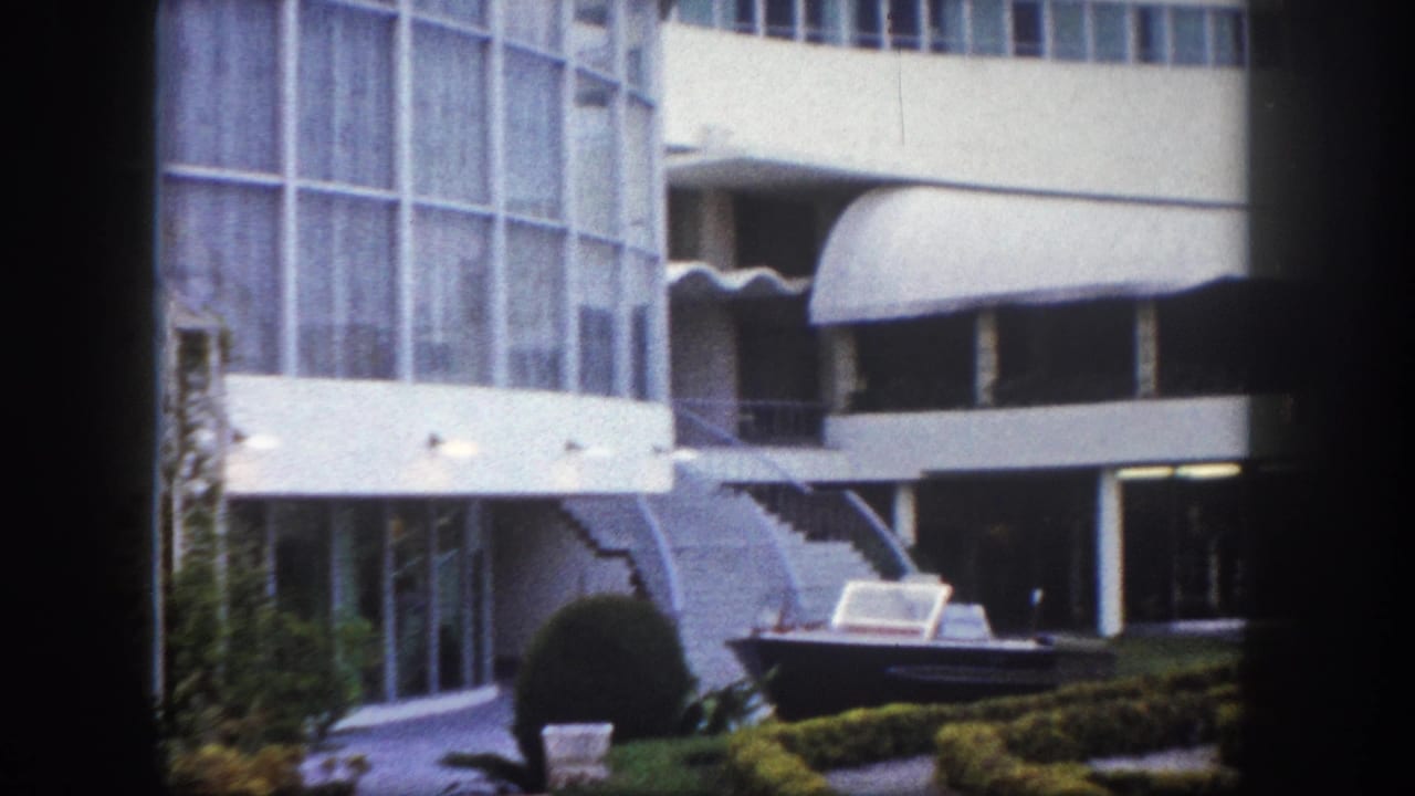 Multi-story modernist hotel exterior with curved canopies, topiary shrubs, and a motorboat on courtyard display, shot on 8mm film.