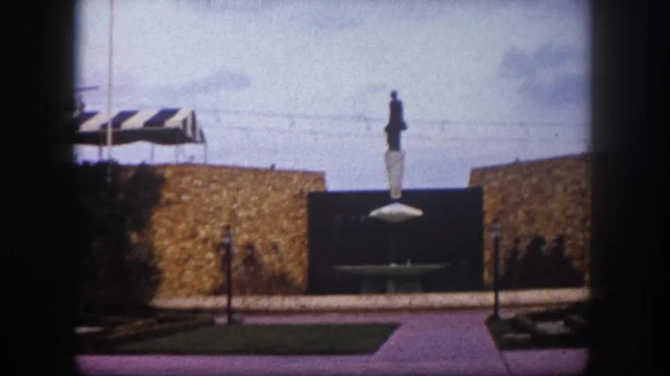 Public monument statue on pedestal, Tampa, Florida, circa 1960 — stone masonry wall, striped canopy, lamp standards, and open plaza in the same frame.