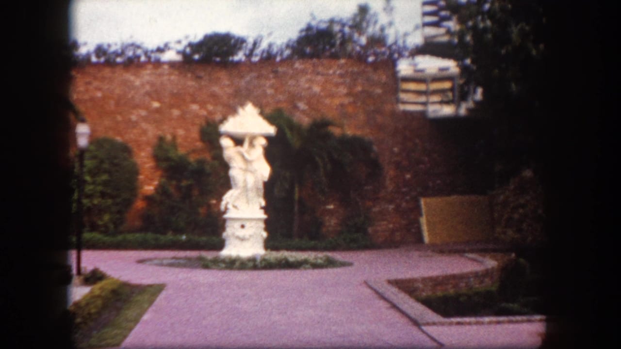 White figurative stone statue on a pedestal in a manicured garden with a red brick wall behind it.