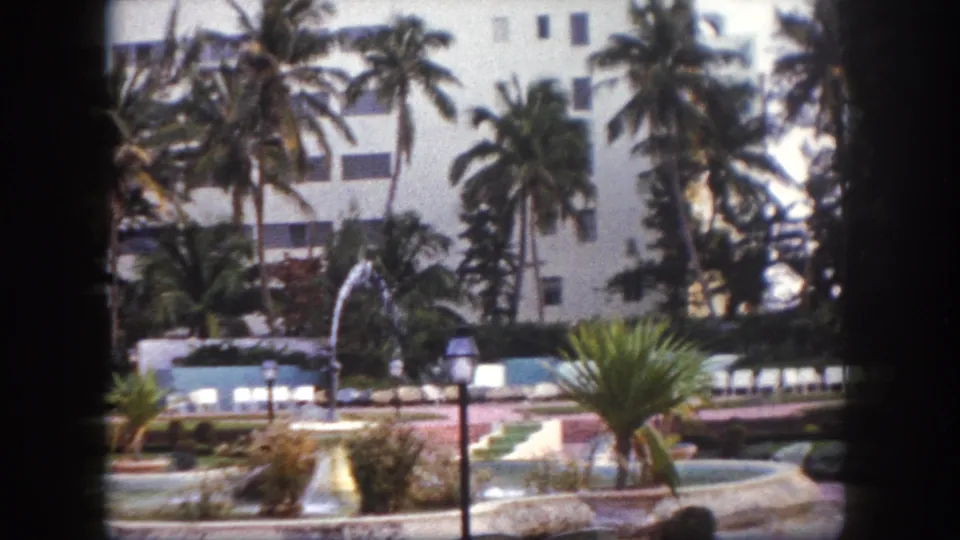 Poolside grounds at a Tampa, Florida resort circa 1960, showing a fountain, cycad palm, lamp posts, and lounge chairs along the pool edge on 8mm film.