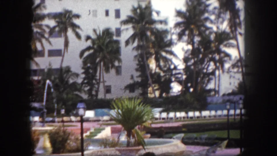Continuing static shot of the poolside fountain area at a Tampa, Florida resort circa 1960, with palm trees, lamp posts, and poolside lounge chairs on 8mm film.
