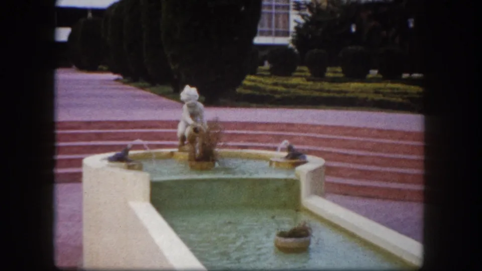 1960s 8mm close-up of an ornamental resort fountain — small figurative statue in a square basin with water jets, red-tiled steps, and clipped hedges, Tampa, Florida area.
