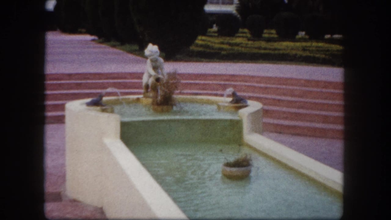 8mm close-up of a decorative fountain with white figurine on pedestal and green-tinted tiered basin, entrance plaza, Tampa, Florida, circa 1960.