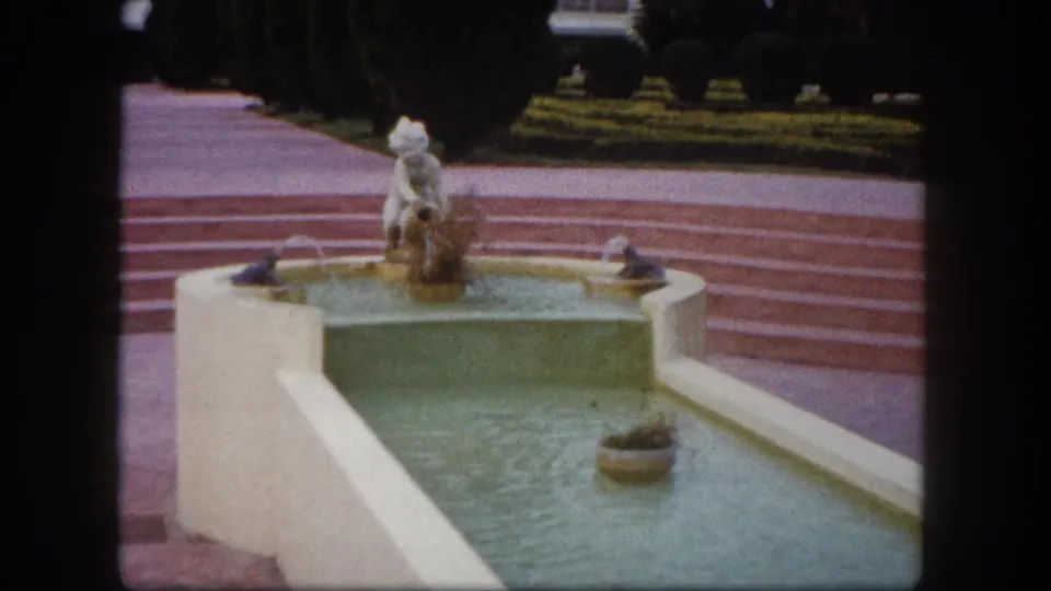 8mm close-up of a decorative fountain with white figurine on pedestal and green-tinted tiered basin, entrance plaza, Tampa, Florida, circa 1960.