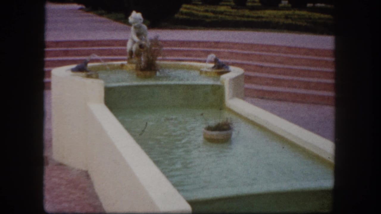 Decorative fountain basin from a slightly wider angle; round urn planter in water, white figurine on pedestal, red-tiled striped steps behind.