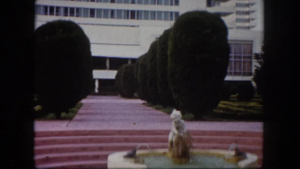 8mm wide shot of a mid-century modernist complex entrance with spherical clipped topiaries, grid-windowed institutional building, and ornamental fountain, Tampa, Florida, circa 1960.