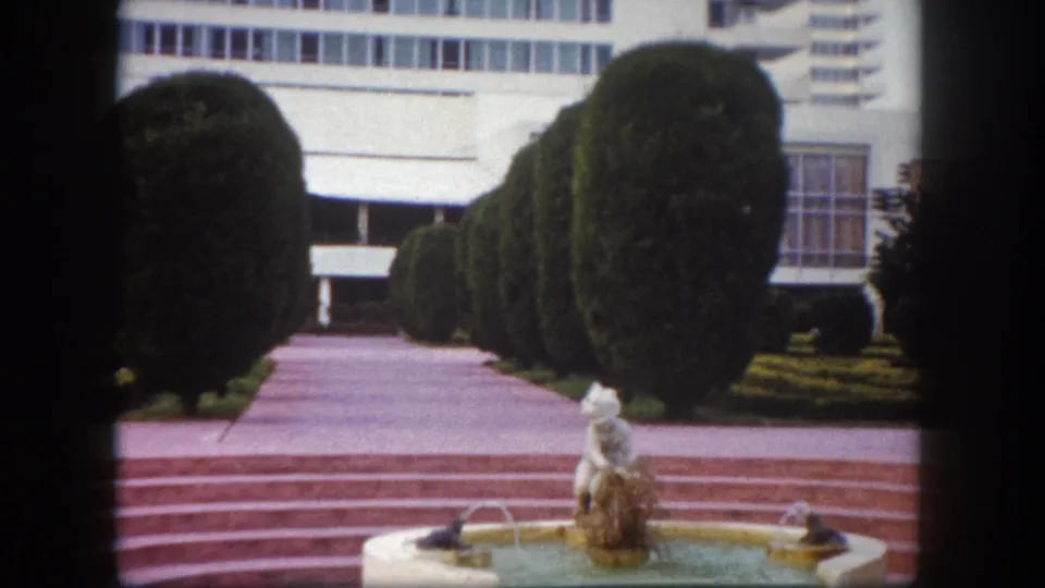 8mm footage of a mid-century complex entrance plaza with rounded topiary hedges, light-colored institutional building facade, and ornamental fountain, Tampa, Florida, circa 1960.
