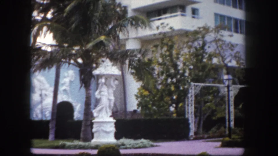 8mm frame circa 1960 showing a classical garden statue before what appears to be a large modernist Florida resort building, with white pergola trellis and palm trees.