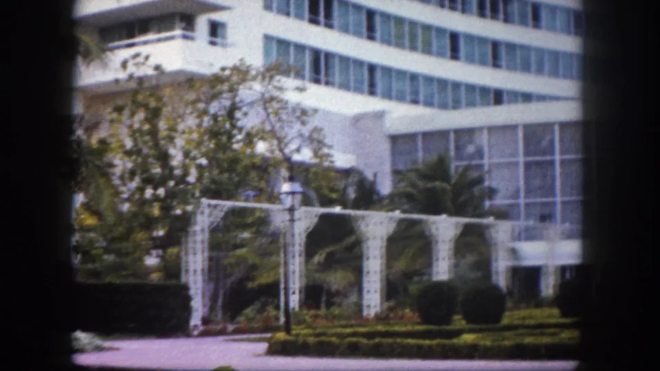 8mm frame circa 1960 showing the exterior of a large modernist Florida building, possibly a resort hotel, with ornamental garden trellis and tropical plantings.