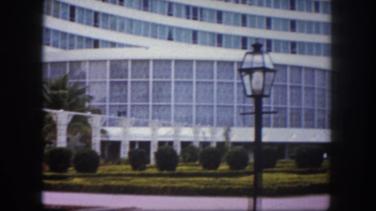 Large curved building with horizontal window banding; prominent period lamppost in right foreground with manicured hedges and topiaries below.