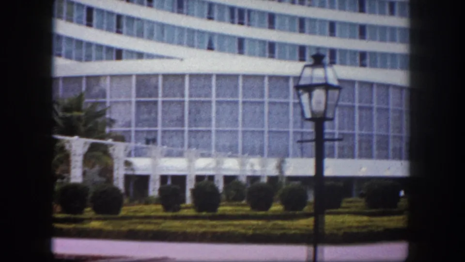 8mm film frame circa 1960, Tampa, Florida: panned view of large curved glass building with formal garden, low hedgerows, and a classic lamppost prominent in the right foreground.