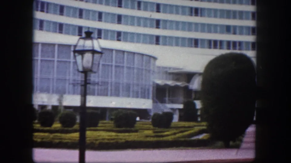 8mm film frame circa 1960, Tampa, Florida: nearer view of curved building entrance with a canopy awning, prominent round topiary shrubs in foreground, and lamppost at left.