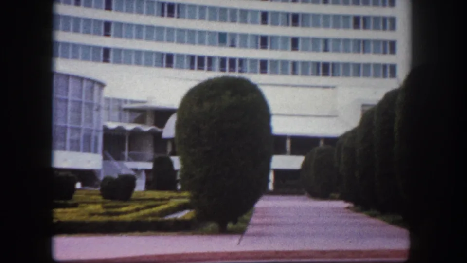 8mm film frame circa 1960, Tampa, Florida: pan continues across formal garden with prominent round topiary shrubs, period lamppost, and curved glass building facade.