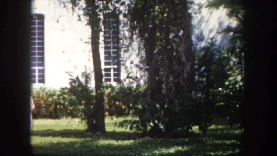 8mm film circa 1960, Tampa Florida — institutional white building, tall multi-paned windows, mature trees, sunlit lawn; one figure just entering frame at right.