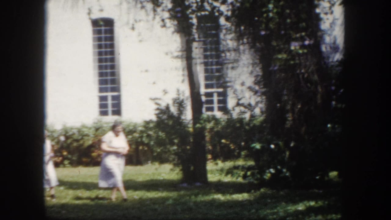 8mm home movie circa 1960, Tampa Florida — elderly woman in light summer dress crossing a sunlit lawn; white-painted building with tall multi-paned windows behind.
