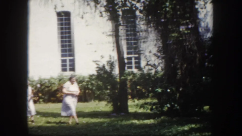 8mm home movie circa 1960, Tampa Florida — elderly woman in light summer dress crossing a sunlit lawn; white-painted building with tall multi-paned windows behind.