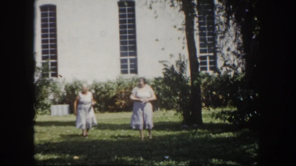 8mm home movie circa 1960, Tampa Florida — two elderly women in light dresses walking toward camera on grass; white institutional building with tall multi-paned windows visible behind them.