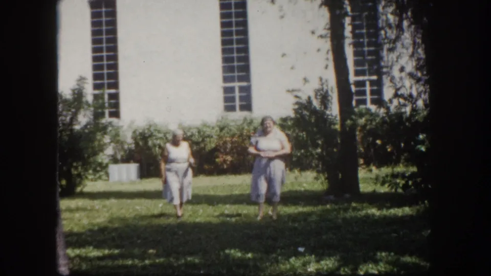 8mm home movie circa 1960, Tampa Florida — two elderly women in light summer attire walking on grass; white building with tall multi-paned windows and trees behind them.