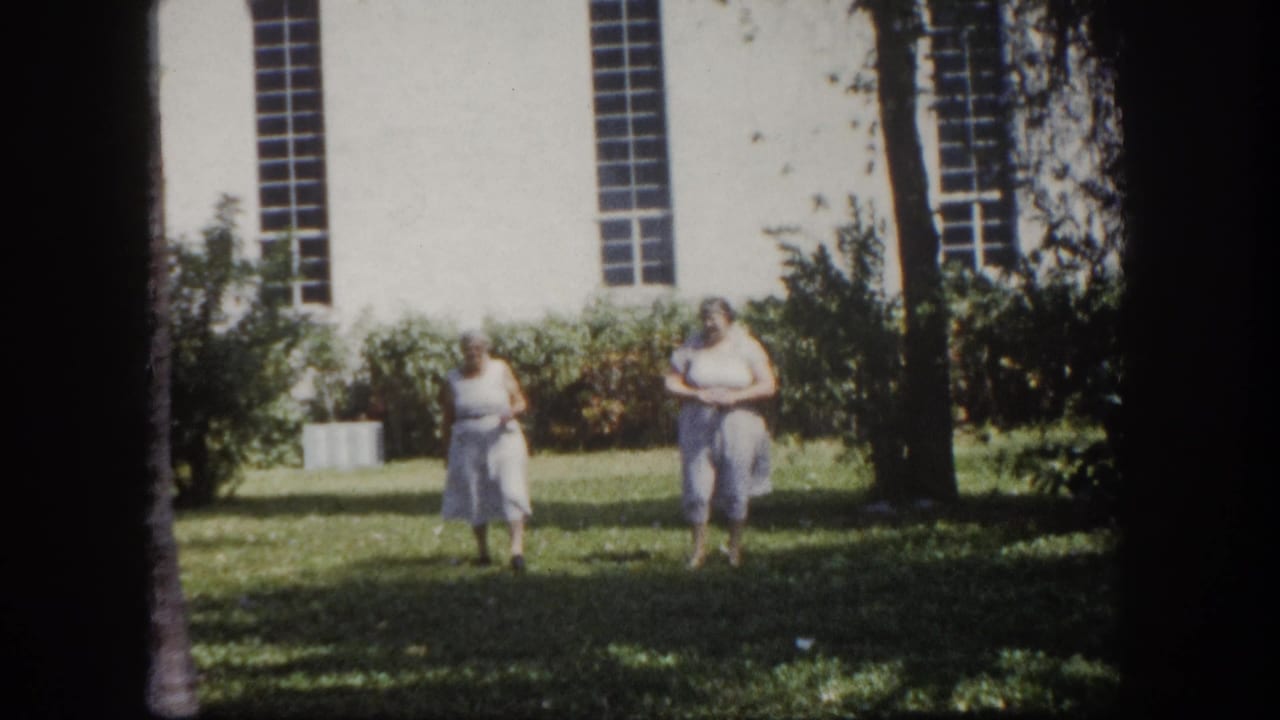 Two elderly women in light summer clothing walking on a green lawn in front of a white building with tall narrow windows.