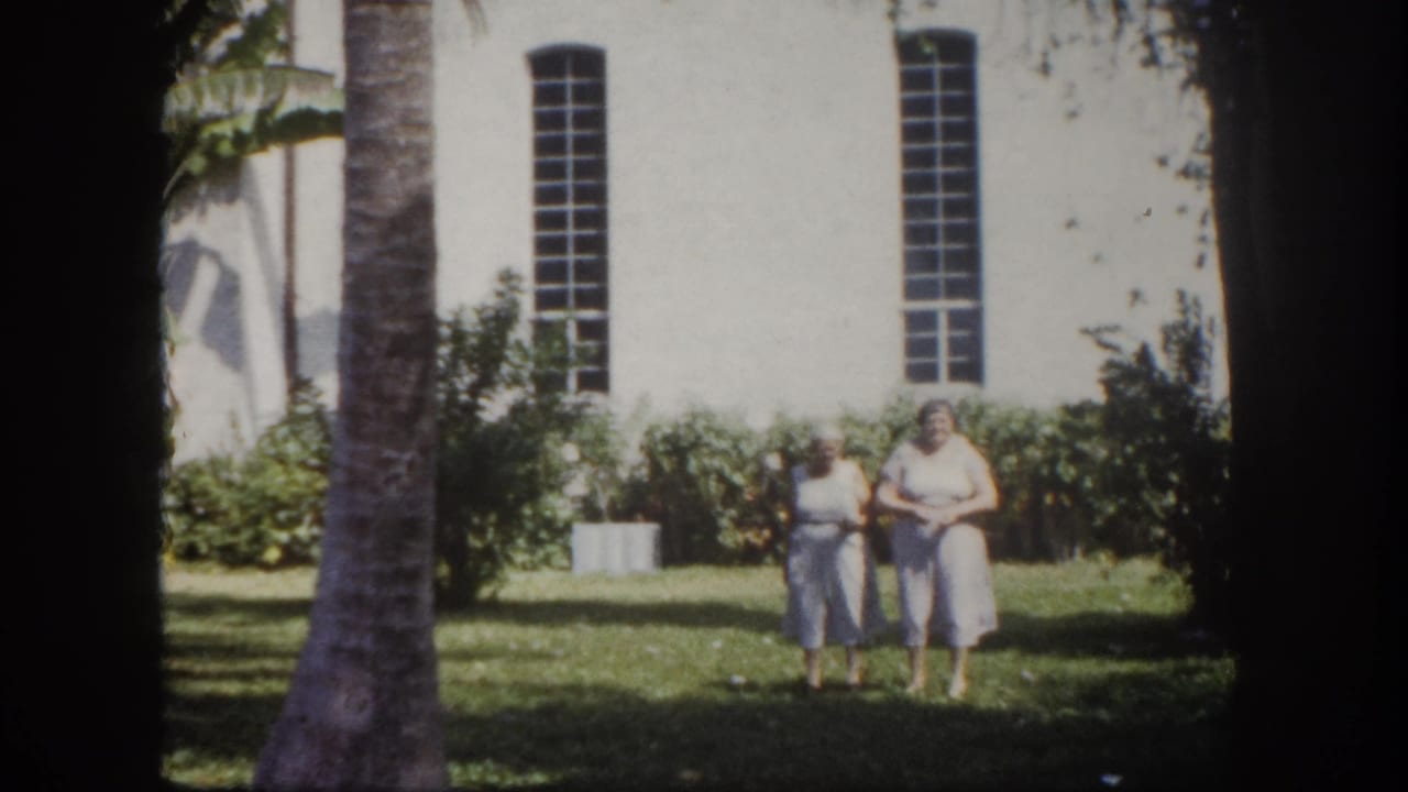 Two women in light-colored clothing walk on a grass lawn in front of a large white building with tall narrow windows.
