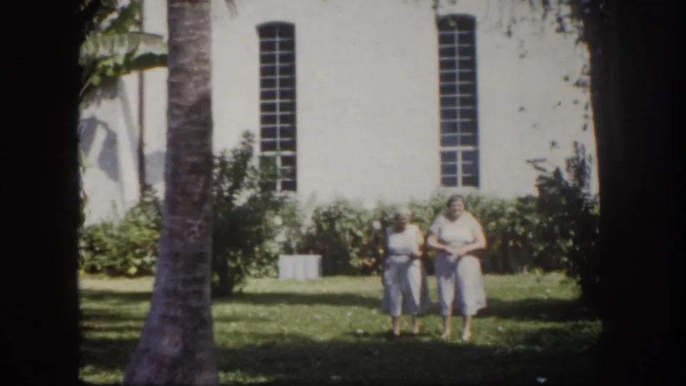 Two adult women in 1960s casual wear walk toward the camera on a sunlit lawn before a white institutional building, Tampa, Florida, circa 1960, 8mm film.