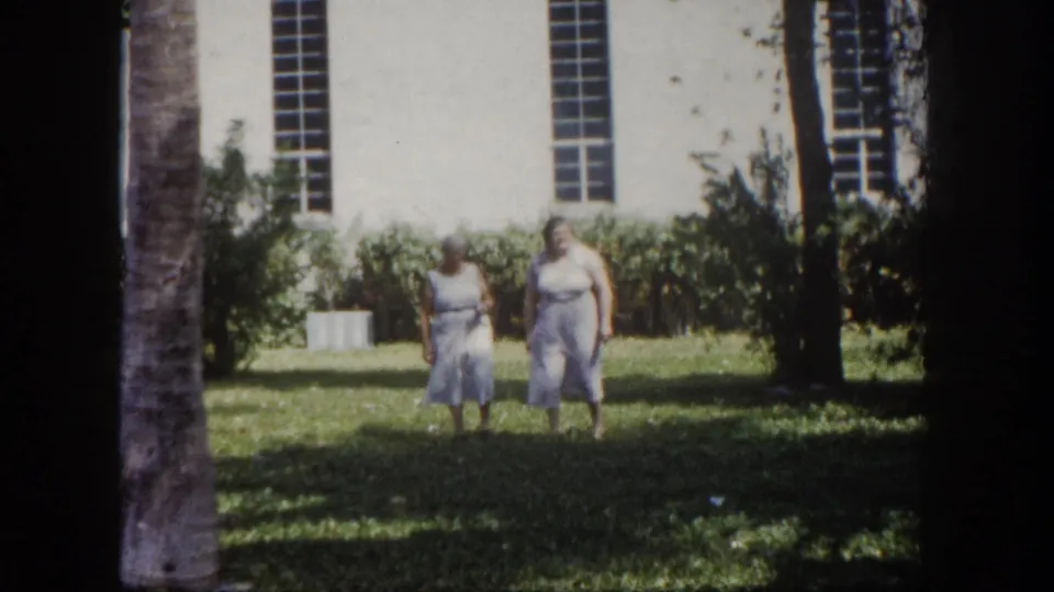 Two adult women pause briefly on the lawn before a white institutional building with tall narrow windows, Tampa, Florida, circa 1960, 8mm film.