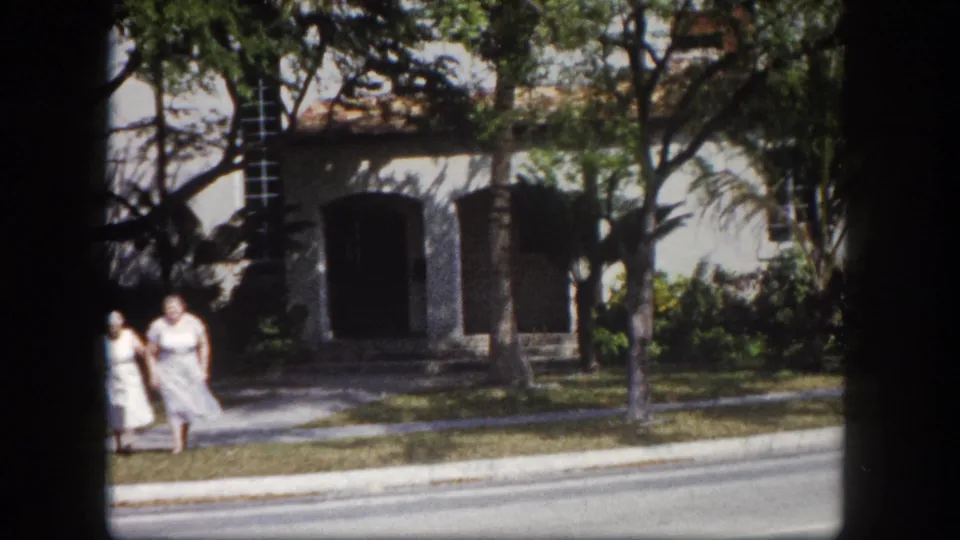 Two women walk past a low stucco building with an arched entrance amid large trees and tropical growth, road in foreground, Tampa, Florida, circa 1960, 8mm.