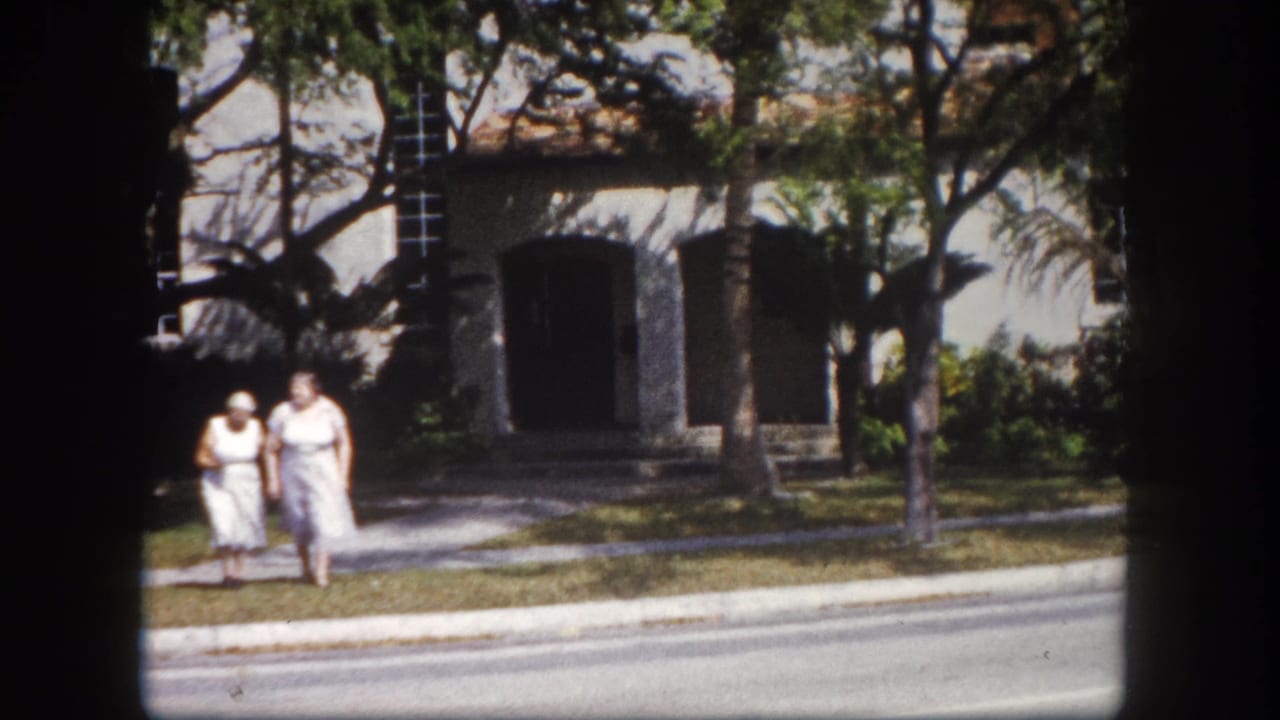 Two adult women in white dresses walk on a sidewalk in Tampa, Florida, circa 1960; stucco building with recessed openings and mature shade trees in background.