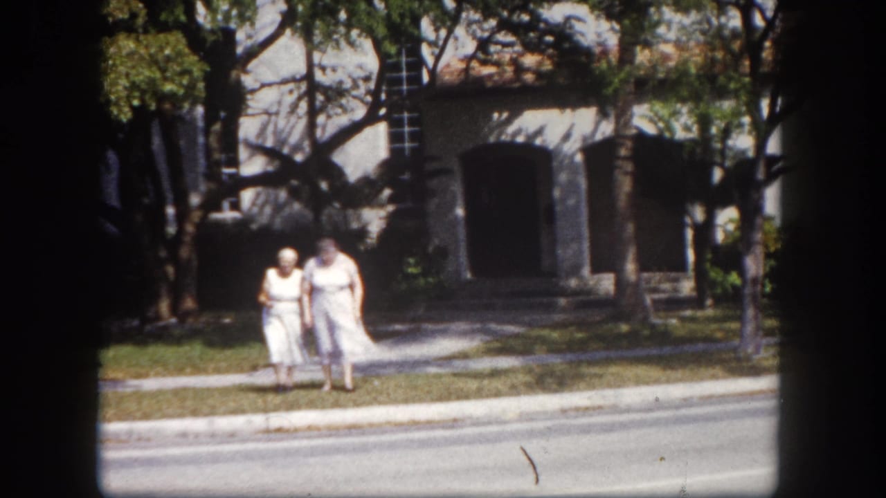 Two women in white dresses walk closer toward the camera; masonry building with dark openings and shade trees behind them.