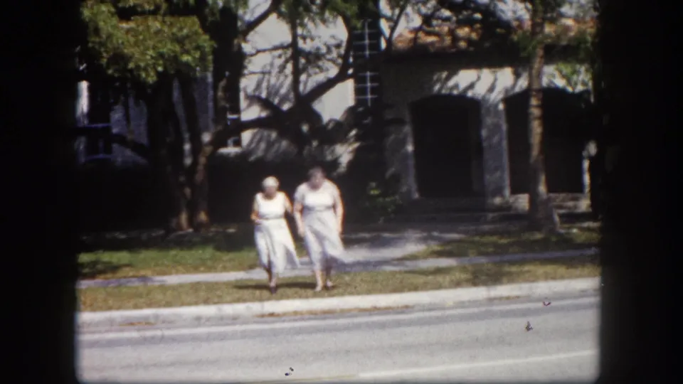 Two adult women in white dresses approach the camera on a Tampa, Florida sidewalk, circa 1960; masonry building and mature trees frame the background.
