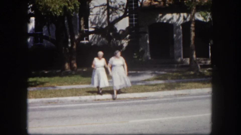 Two adult women in white dresses near the camera on a Tampa, Florida sidewalk, circa 1960; faces clearly visible, stucco building with dark arched openings behind them.