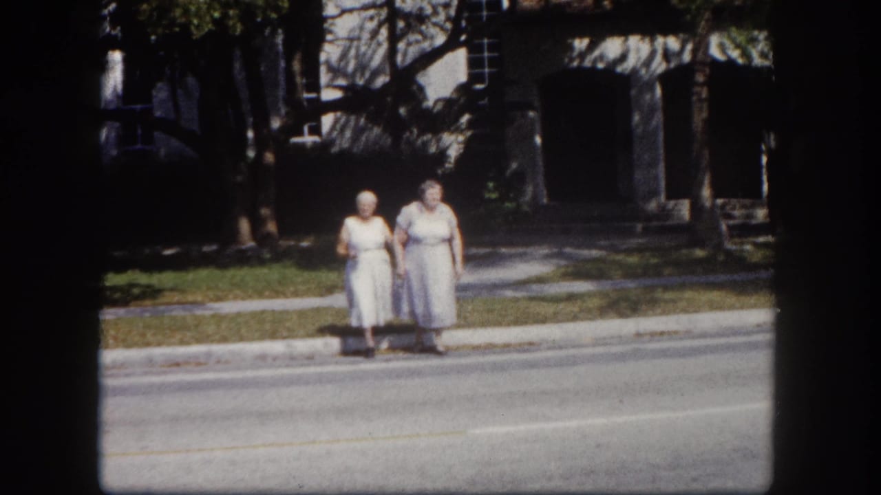 Two elderly women in light dresses standing at a roadside curb, trees and a building visible behind them.