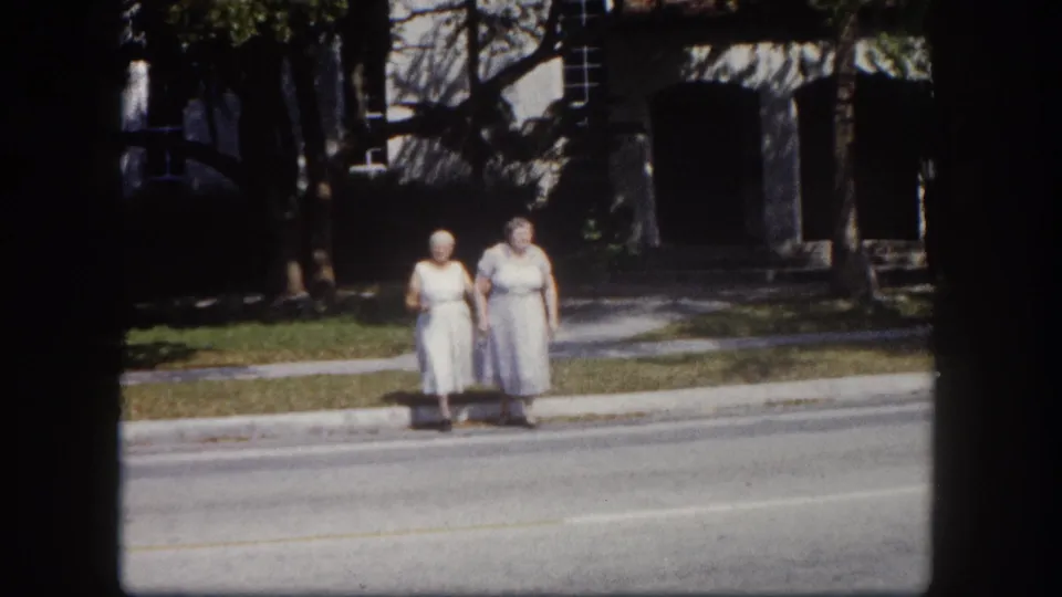 Two elderly women in light mid-length dresses stand at the edge of a sunny street in what appears to be a residential Tampa, Florida neighborhood, circa 1960, 8mm film.