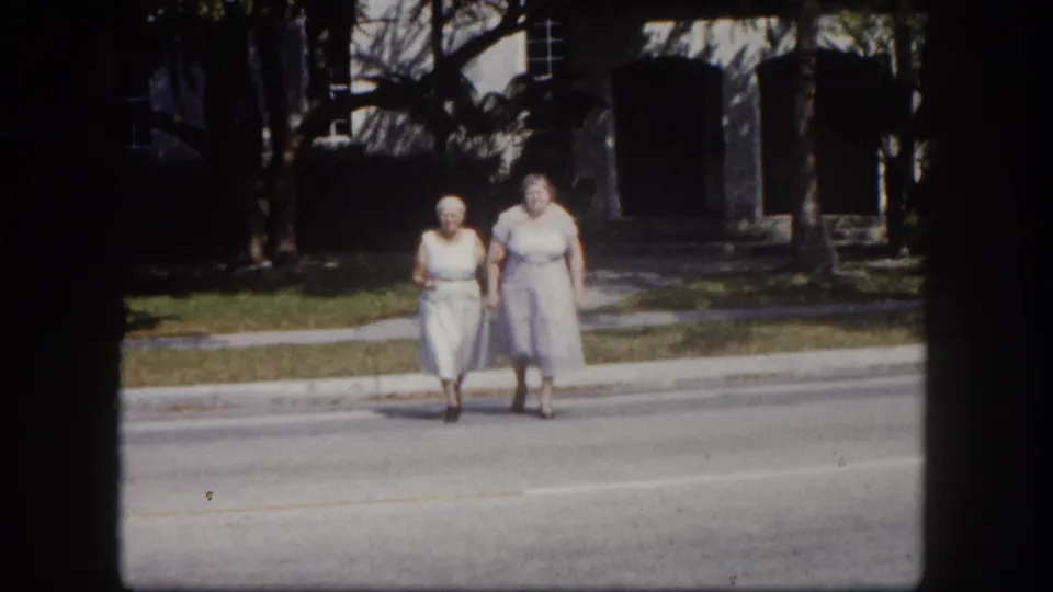 Two elderly women walk side by side toward the camera across a quiet street, tropical vegetation and a masonry building visible behind — 8mm Florida home movie, circa 1960.