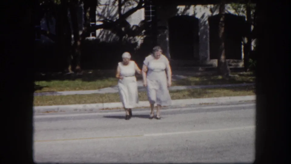 Two elderly women approach the camera across a residential Tampa, Florida street circa 1960, their faces visible in this 8mm home movie frame.