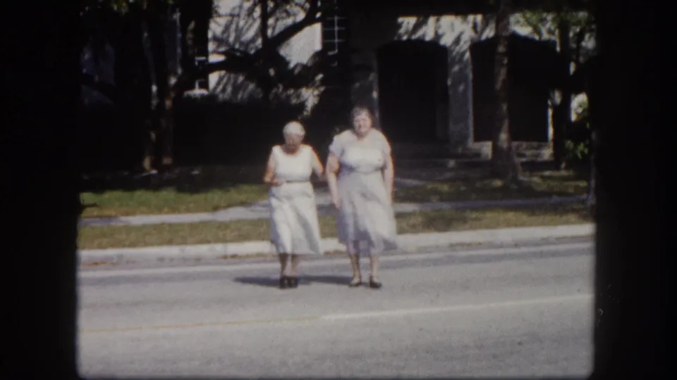 Two elderly women in early-1960s pale dresses cross a quiet residential street, likely Tampa, Florida, circa 1960, filmed on 8mm color stock.