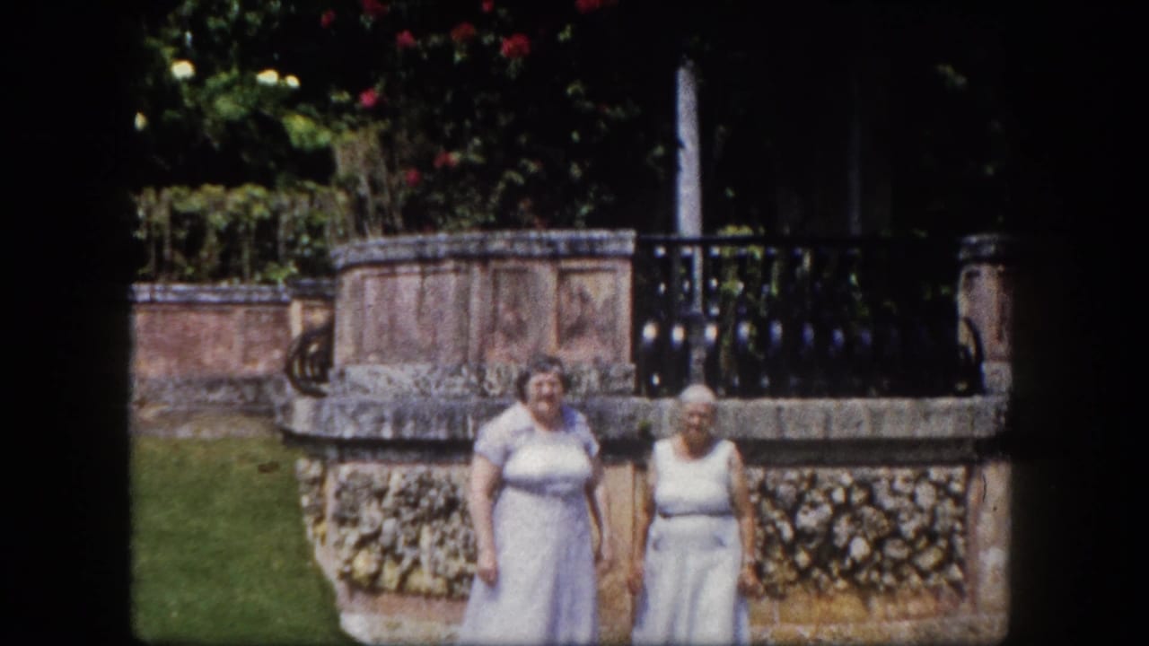 Two elderly women in light dresses stand before a coral-stone garden wall topped with black iron railing; red-flowering shrubs rise behind them.