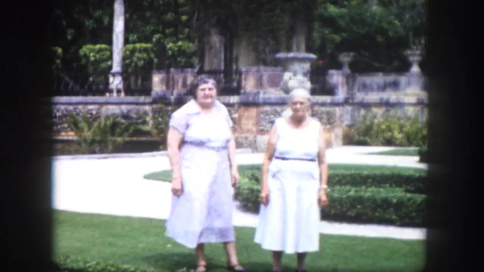 8mm home movie circa 1960, Tampa Florida — two older women in light sleeveless dresses posed on a formal garden lawn, with ornamental stone arched colonnade and trimmed hedges behind them.