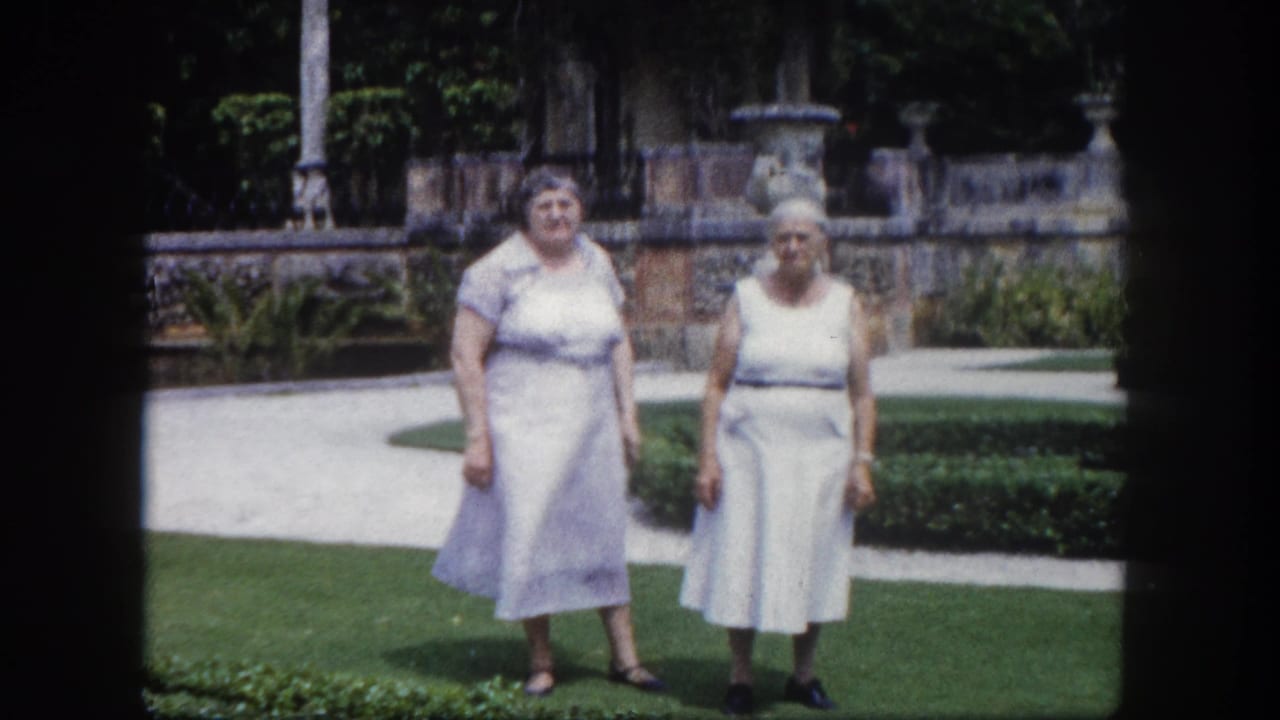 Two women in pale light-colored dresses standing on a manicured lawn in a formal garden with stone urns and trimmed hedges behind them.